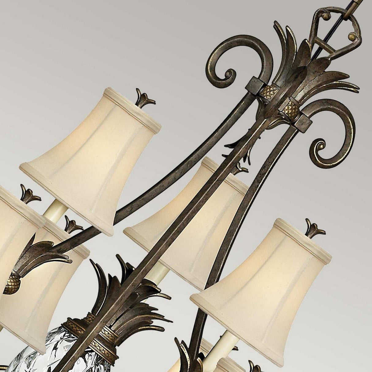 Close-up of the Plantation 10 Light Chandelier in bronze, showcasing cream-colored fabric lampshades and intricate metalwork with decorative cast detailing against a light gray background. The design includes filigree and pineapple motifs.