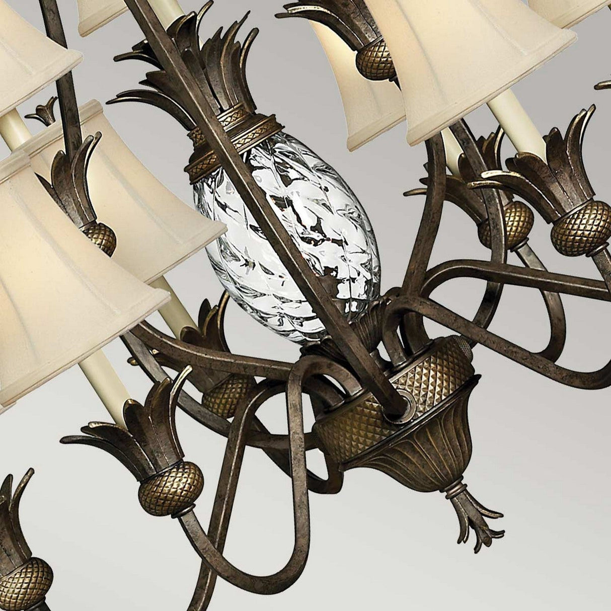 The Plantation 10 Light Chandelier - Bronze is displayed in close-up, highlighting its intricately detailed vintage bronze finish. It features several cream-colored lampshades and elegant pineapple motifs encircling a clear, textured glass centerpiece against a plain light gray background.