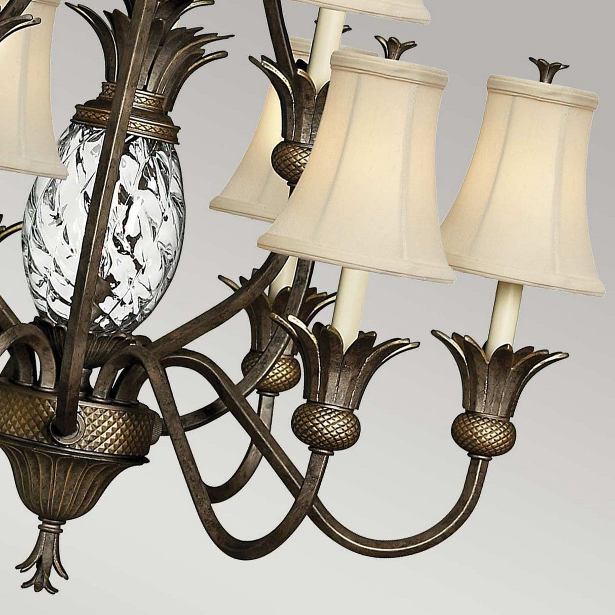 The Plantation 10 Light Chandelier in bronze boasts decorative cast detailing and six elegant cream-colored lampshades. Its central fixture is adorned with a pineapple-shaped glass element featuring swirling patterns, creating a classic and elegant style against a neutral background.