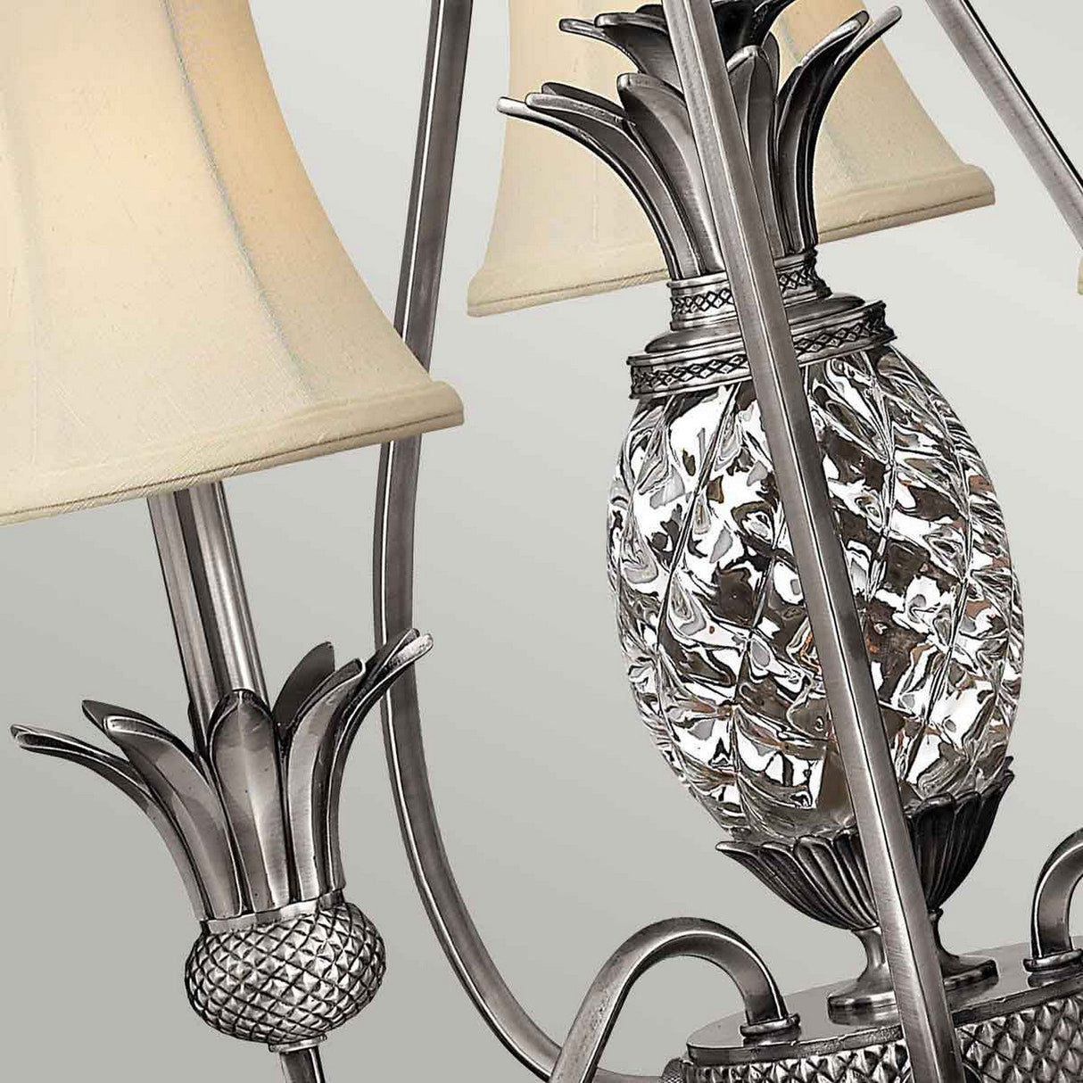 Close-up of the Plantation 3 Light Chandelier, featuring decorative metal work with pineapple motifs and beige fabric lampshades. The intricate design includes textured leaves and clear optic glass details, set against a soft gray background, capturing the elegance of the Plantation collection in its polished antique nickel finish.