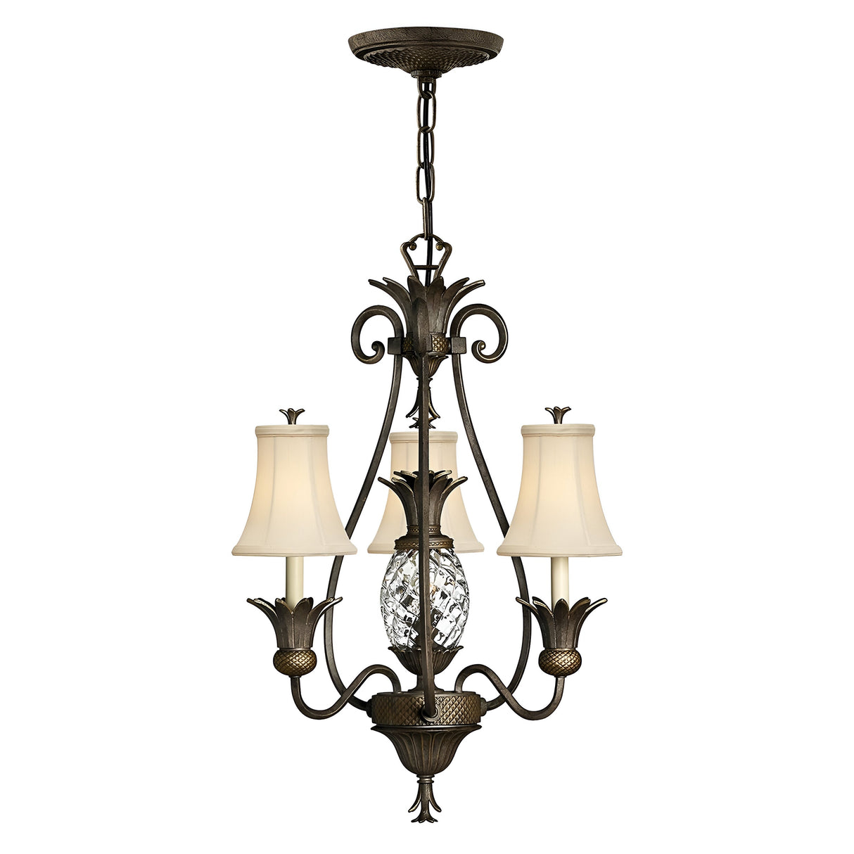 The Plantation 3 Light Chandelier in bronze showcases three cream-colored fabric lampshades adorned with intricate scrollwork, while a clear optic glass pineapple design graces the central structure for an elegant and classic touch.