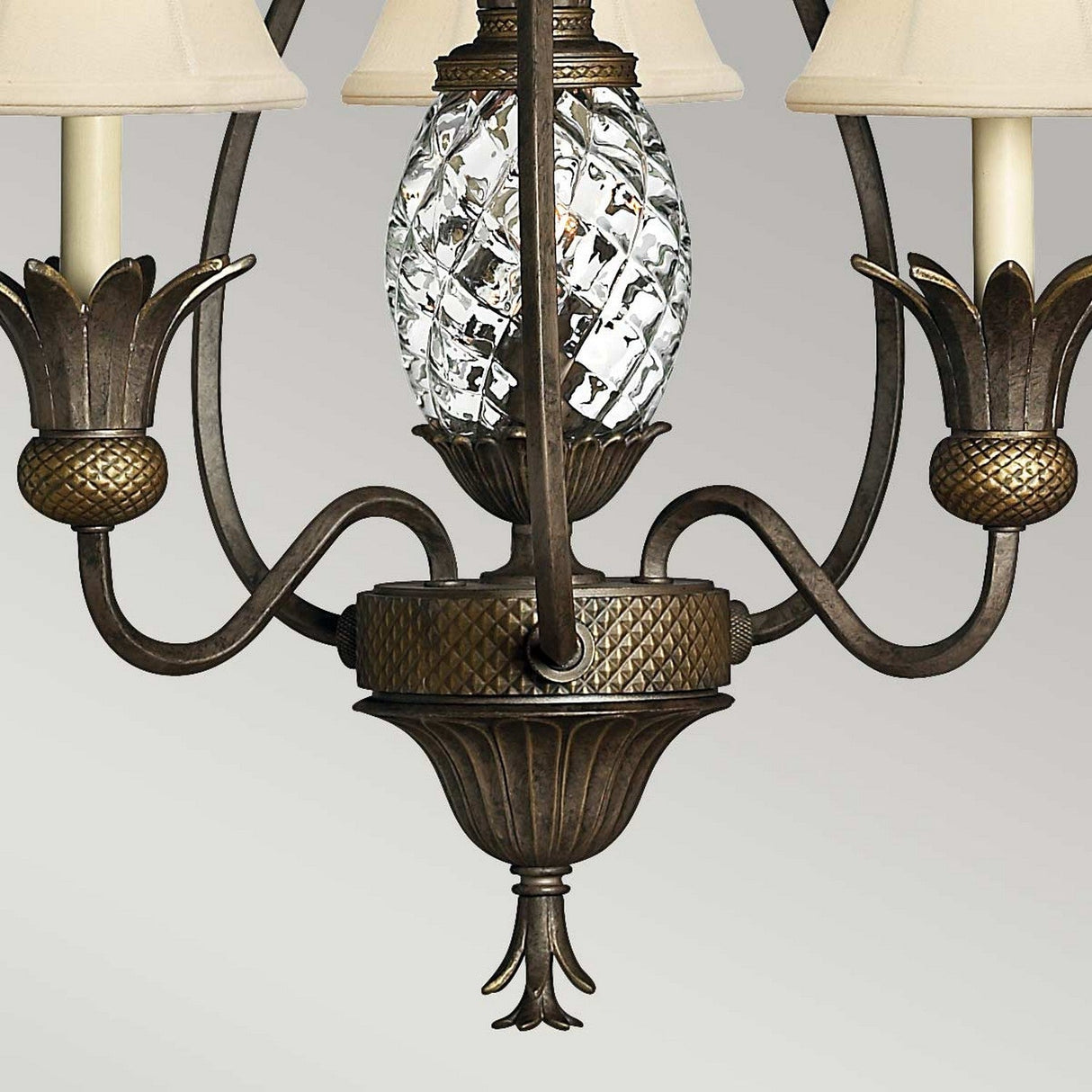 The Plantation 3 Light Chandelier - Bronze showcases an exquisite central crystal pineapple design, complemented by intricate patterns and three candle-style arms adorned with small bronze pineapples and beige lampshades, all in a stunning Pearl Bronze finish.