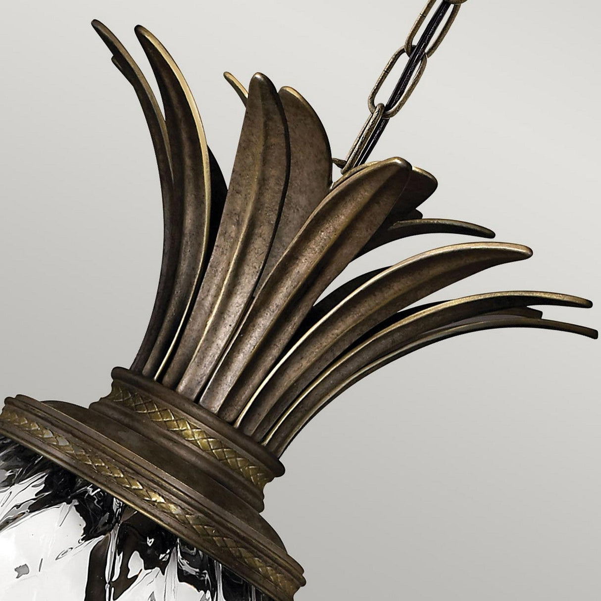 This close-up highlights the Plantation 4 Light Pendant - Bronze, showcasing its antique bronze finish adorned with palm frond details. The fixture features a pineapple-shaped optic glass that adds elegance and hangs gracefully from a metal chain against a simple, light gray background.