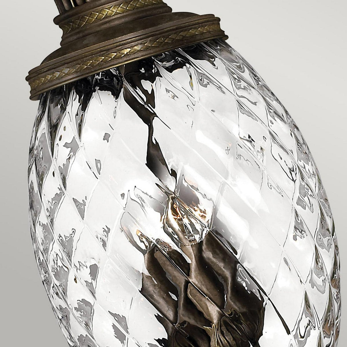The close-up showcases the Plantation 4 Light Pendant - Bronze, which features decorative optic glass with a crisscross pattern. Its bronze-like metal fittings elegantly top the pineapple-shaped transparent glass, revealing the filament inside. The neutral gray backdrop perfectly complements its vintage charm.