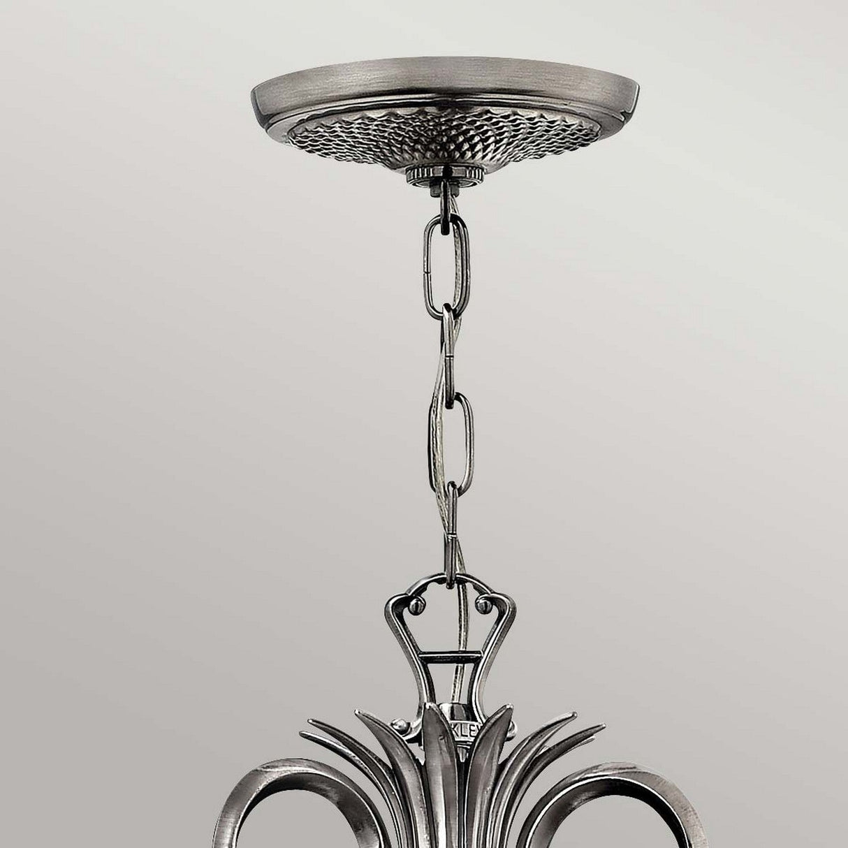 A close-up of the Plantation 7 Light Chandelier in polished antique nickel showcases its ornate design, with a chain connecting the decorative base to an intricately designed hanging element. The chandelier is adorned with curved accents and pineapple-shaped clear optic glass, set against a neutral background.