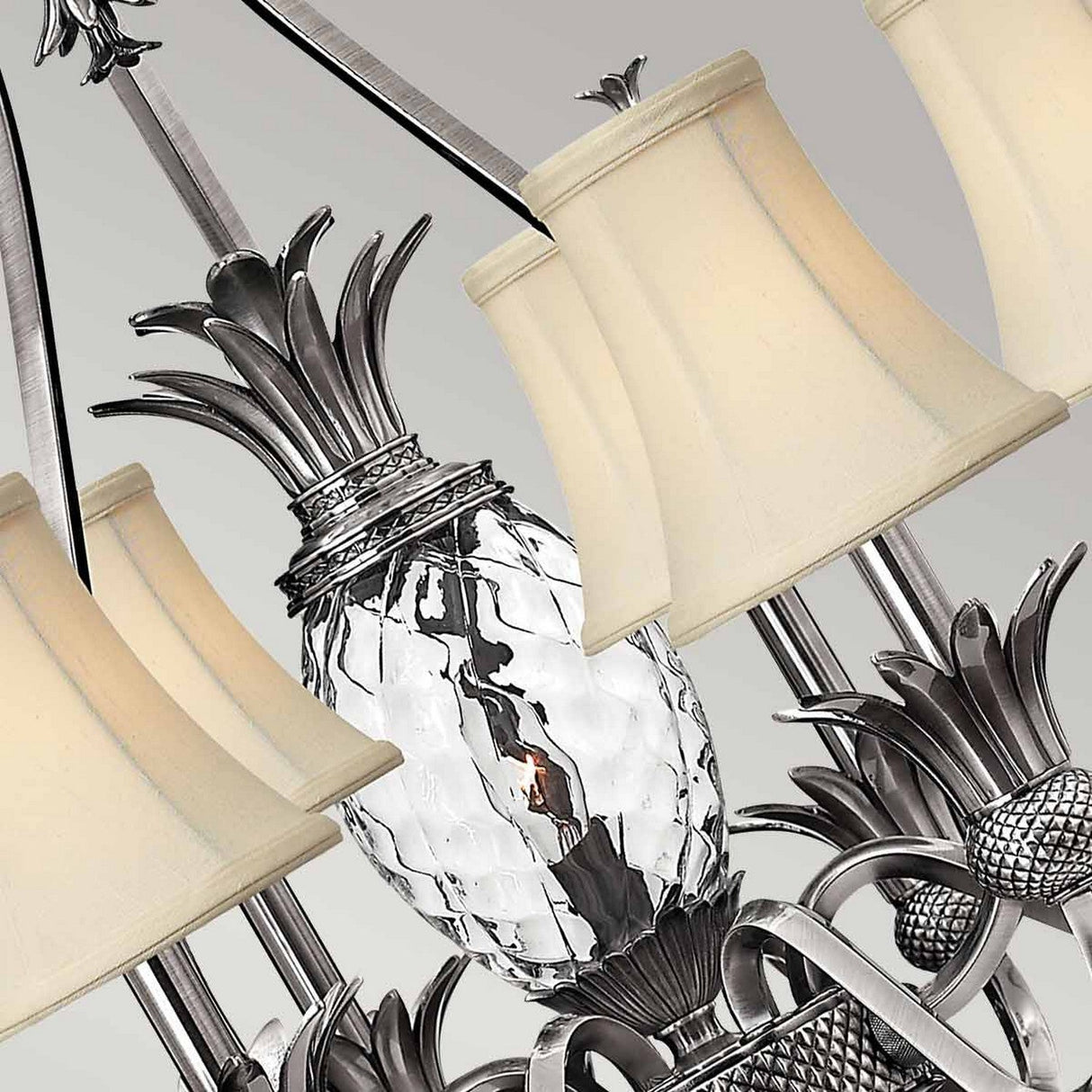 The Plantation 7 Light Chandelier - Polished Antique Nickel features a close-up view highlighting pineapple-shaped clear optic glass elements and multiple cream-colored lampshades set against a neutral background. Its intricate metal frame, crafted in polished antique nickel, exudes timeless elegance.