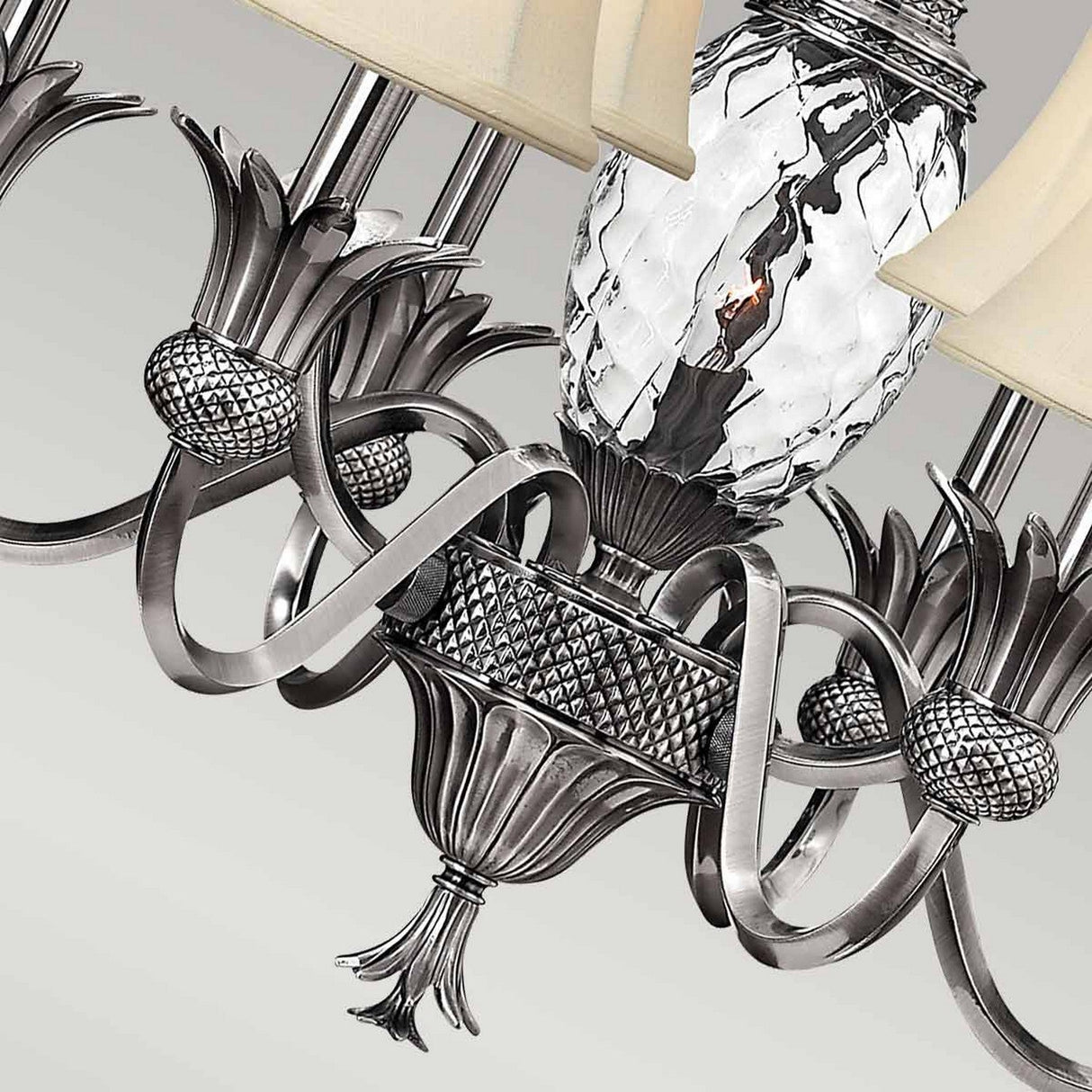 Close-up of the Plantation 7 Light Chandelier - Polished Antique Nickel, showcasing its ornate metalwork with intricate floral designs and a centerpiece crafted from pineapple-shaped clear optic glass. The chandelier features multiple bulbs adorned with beige lampshades, reflecting an elegant and classic style.