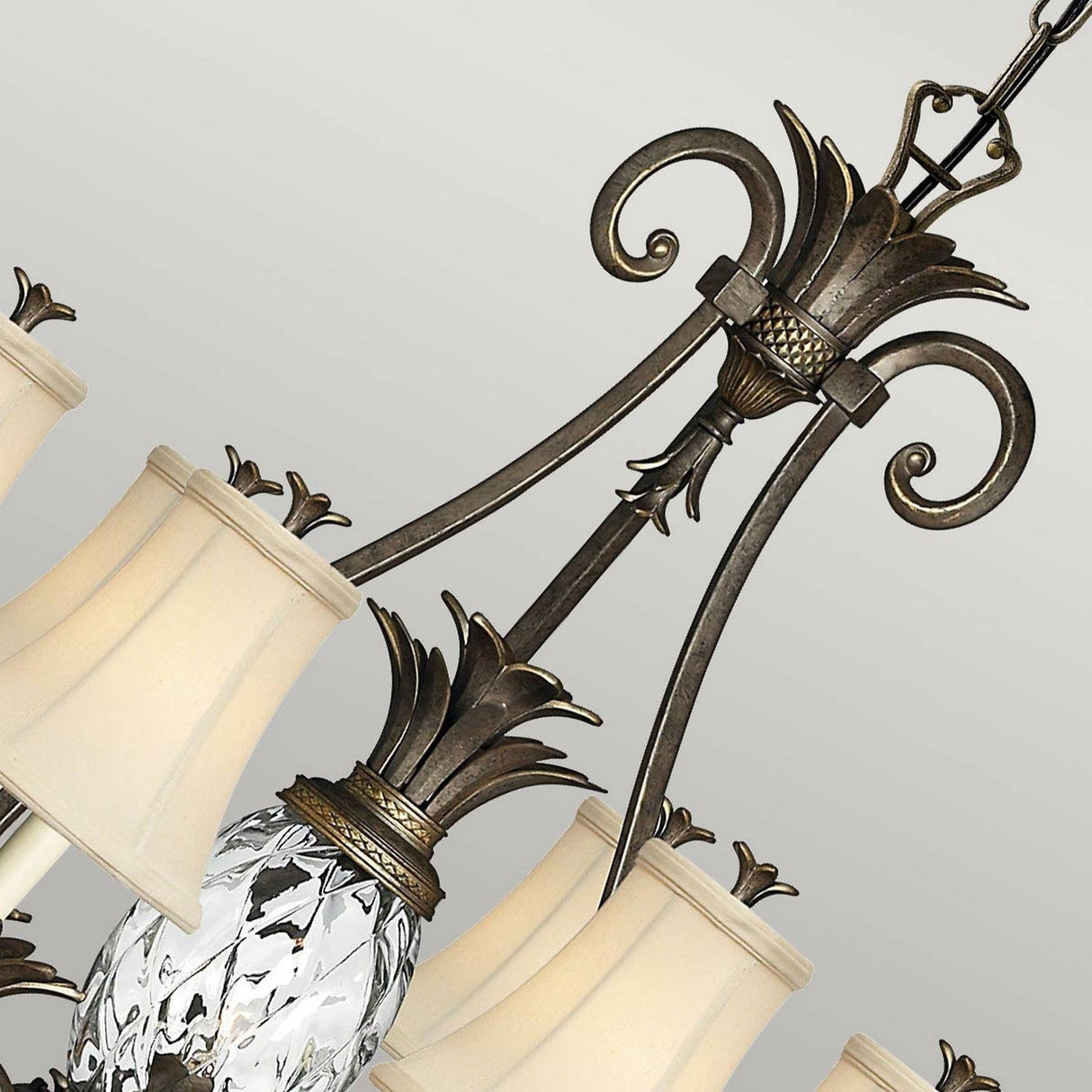 The Plantation 7 Light Chandelier - Pearl Bronze exudes elegance with its intricate metalwork and cream fabric lampshades. It features ornate scroll designs paired with a central pineapple-shaped clear optic glass element, all beautifully highlighted by a stunning Pearl Bronze finish set against a gray background.