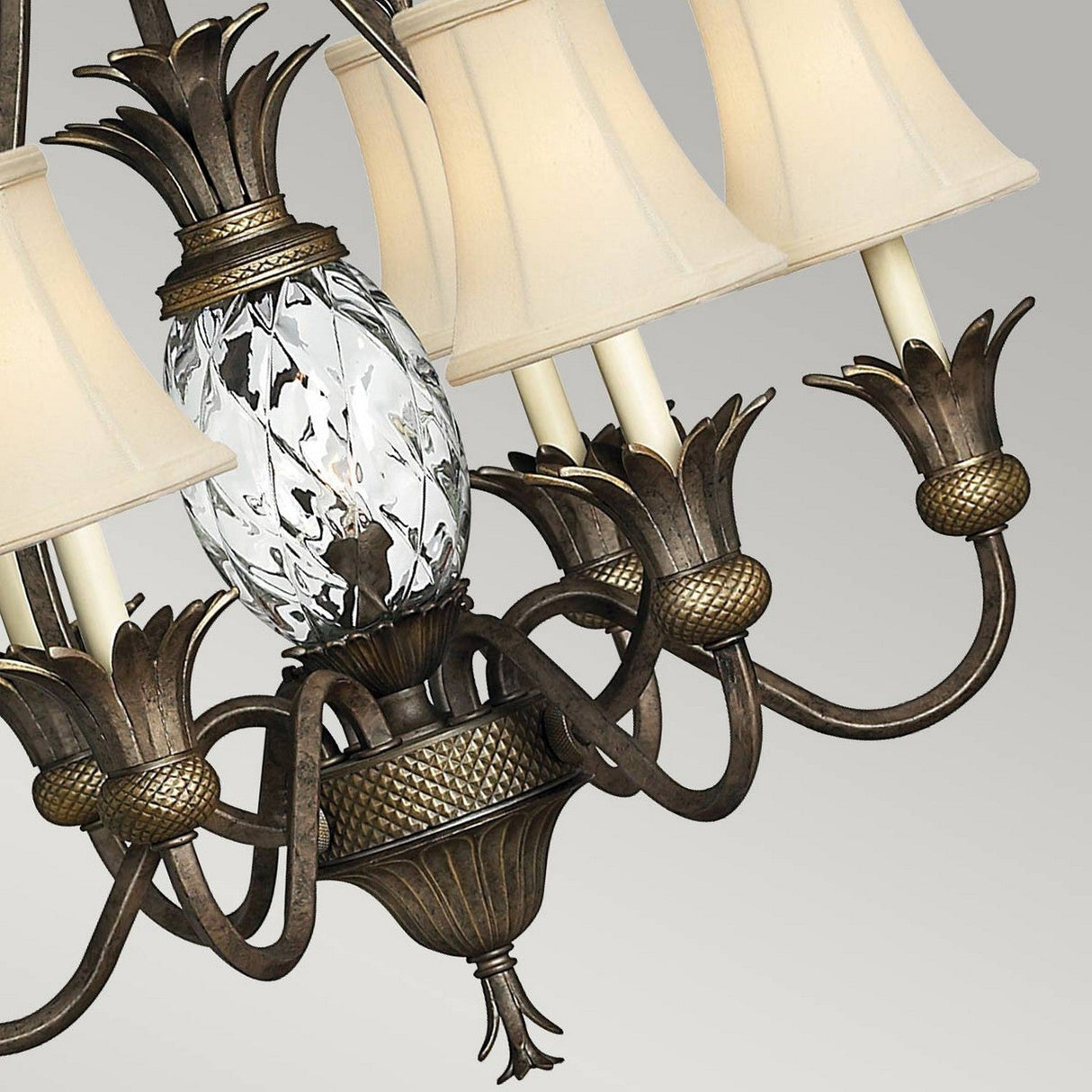 A close-up view of the Plantation 7 Light Chandelier - Pearl Bronze reveals a central clear optic glass fixture in the shape of an ornate pineapple, adorned with intricate designs. Encircling it are arms finished in pearl bronze, each supporting a lamp with a cream-colored conical shade. The chandelier radiates an elegant, vintage charm against a simple backdrop.