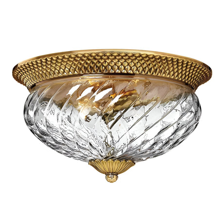 The Plantation 3 Light Flush in Burnished Brass is a ceiling light fixture featuring an elegant combination of gold and clear optic glass with a decorative, textured swirl design. The burnished brass trim boasts intricate detailing and mounts flush against the ceiling for a sophisticated look.