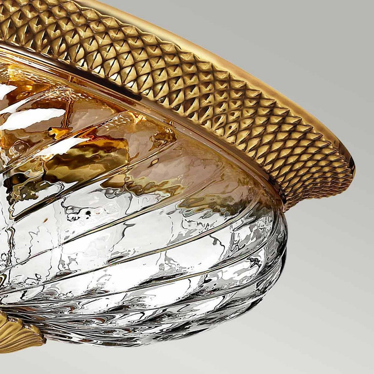 A close-up of the Plantation 3 Light Flush - Burnished Brass fixture highlights its swirling optic glass design and elegant burnished brass rim. The light reflects off the intricate patterns, lending a luxurious appearance against the neutral background.