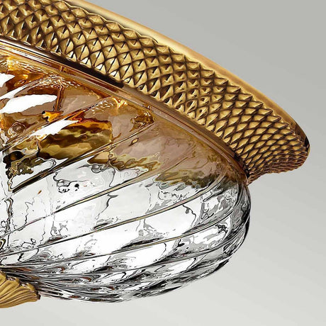 A close-up of the Plantation 3 Light Flush - Burnished Brass fixture highlights its swirling optic glass design and elegant burnished brass rim. The light reflects off the intricate patterns, lending a luxurious appearance against the neutral background.