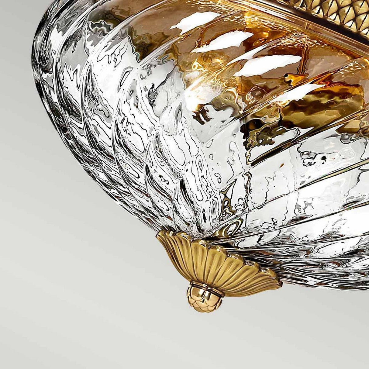 Close-up of the Plantation 3 Light Flush - Burnished Brass ceiling fixture, showcasing ornate swirling designs. The optic glass displays clear spiral textures, complemented by burnished brass accents that impart a vintage and elegant appearance while reflecting warm light beautifully.