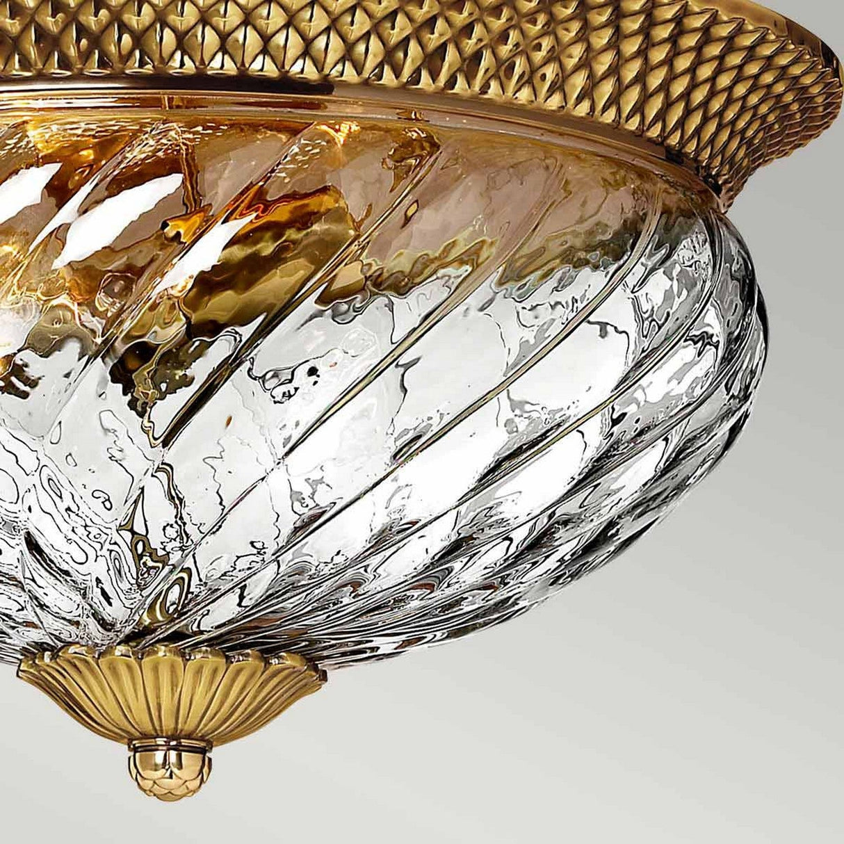 A detailed view of the Plantation 3 Light Flush - Burnished Brass ceiling fixture showcases its sophisticated base with intricate diamond patterns. The optic glass shade, highlighted by swirling clear and gold hues, adds to its ornate appearance.