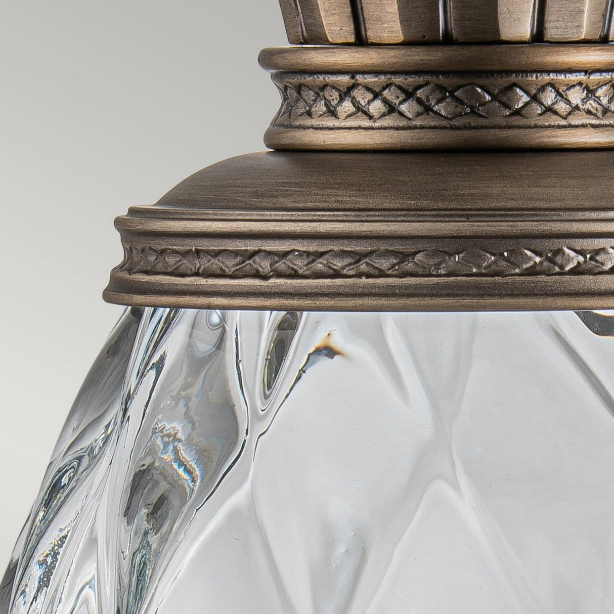A close-up of the Plantation Outdoor 1 Light Medium Wall Lantern reveals its vintage charm, featuring pineapple optic glass and ornate metal trim. The burnished bronze finish highlights intricate detailing, and the unique rippled design enhances its elegance against a light, neutral background.