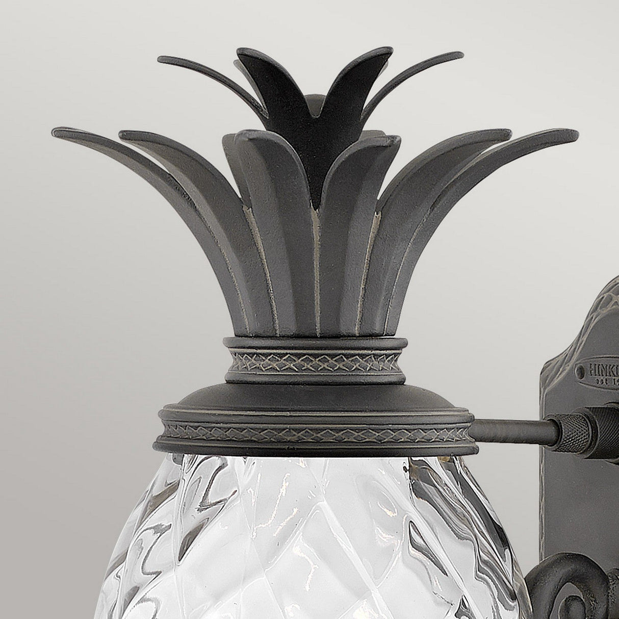 The Plantation Outdoor 1 Light Medium Wall Lantern in black showcases a striking bronze pineapple design adorned with intricately detailed leaves at the top. Its pineapple-shaped glass, textured with a diamond pattern that mimics the surface of a real pineapple, makes this lantern an impressive addition to any outdoor lighting setup.