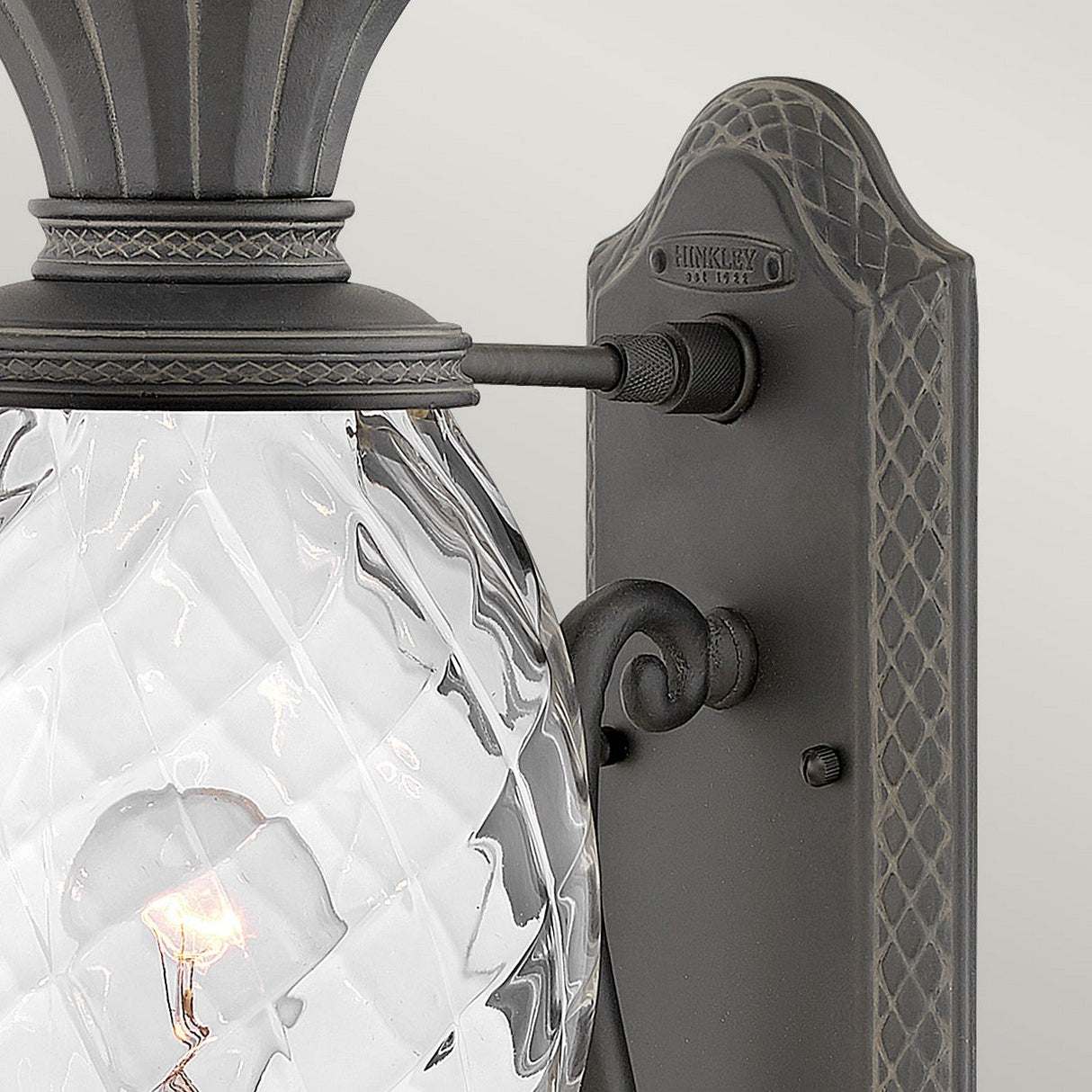A close-up view of the Plantation Outdoor 1 Light Medium Wall Lantern - Black showcases its vintage-style design with a dark metal frame. The lantern features a textured, diamond-patterned, pineapple-shaped glass cover that partially reveals the bulb inside. A decorative arm elegantly connects this outdoor lighting fixture to a patterned backplate.