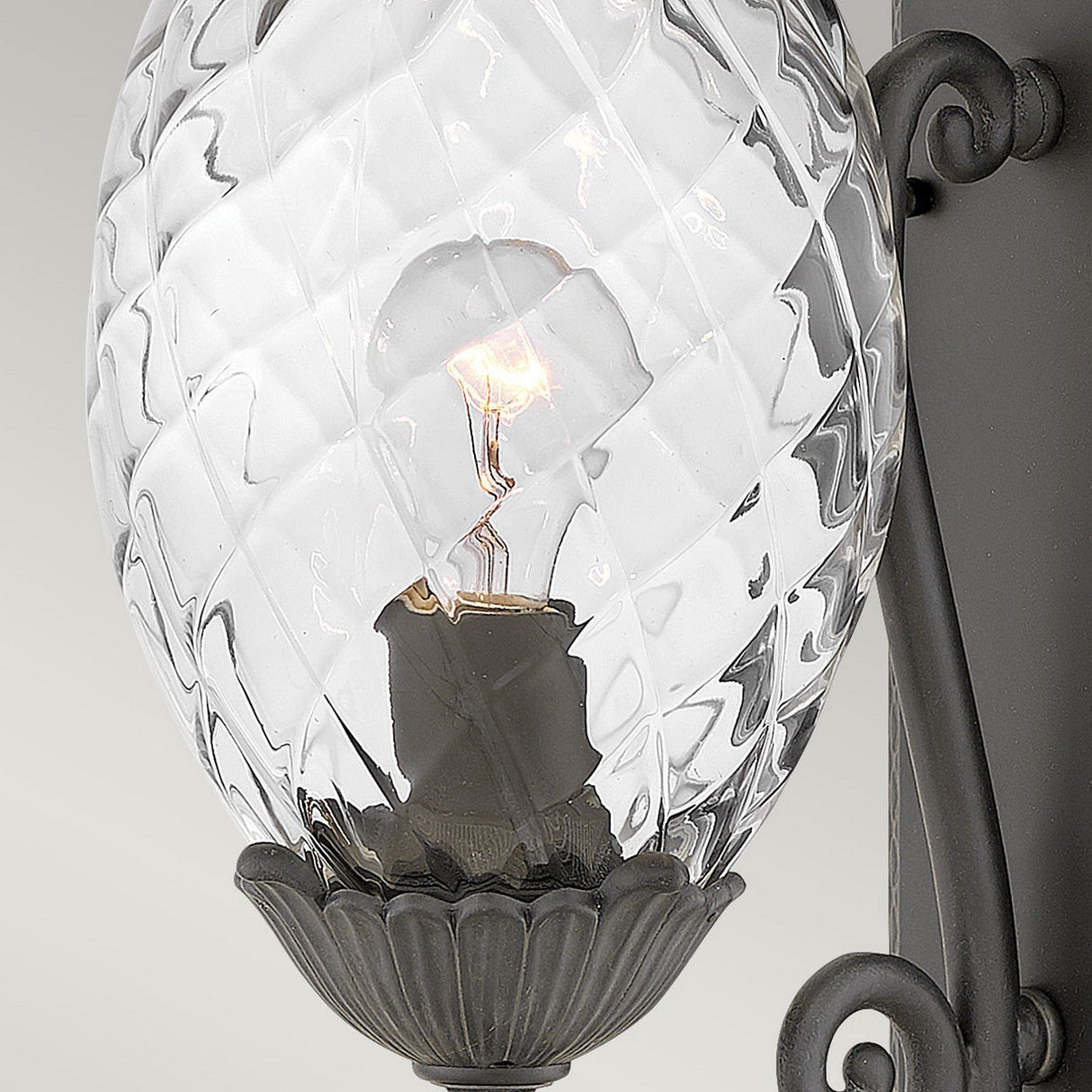 A close-up of the Plantation Outdoor 1 Light Medium Wall Lantern in black showcases its decorative pineapple-shaped glass shade. A glowing bulb is visible inside, surrounded by intricate black metalwork, making it a perfect choice for outdoor lighting.