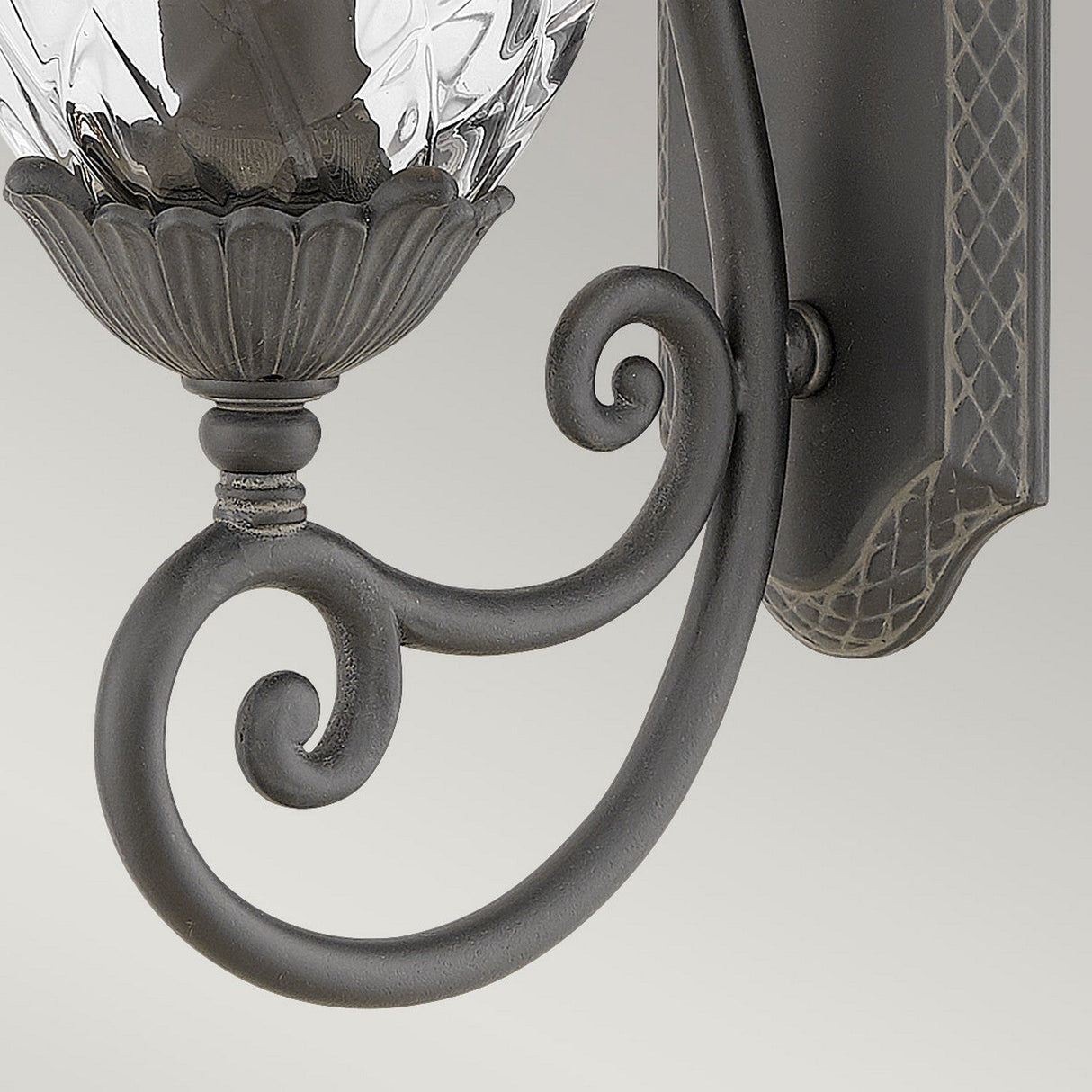 Close-up of the Plantation Outdoor 1 Light Medium Wall Lantern in black, showcasing its decorative metal design with intricate scrollwork and a pineapple-shaped glass lampshade featuring a textured pattern. The backdrop is a plain, soft gray color, ideal for blending indoor elegance with outdoor lighting styles.