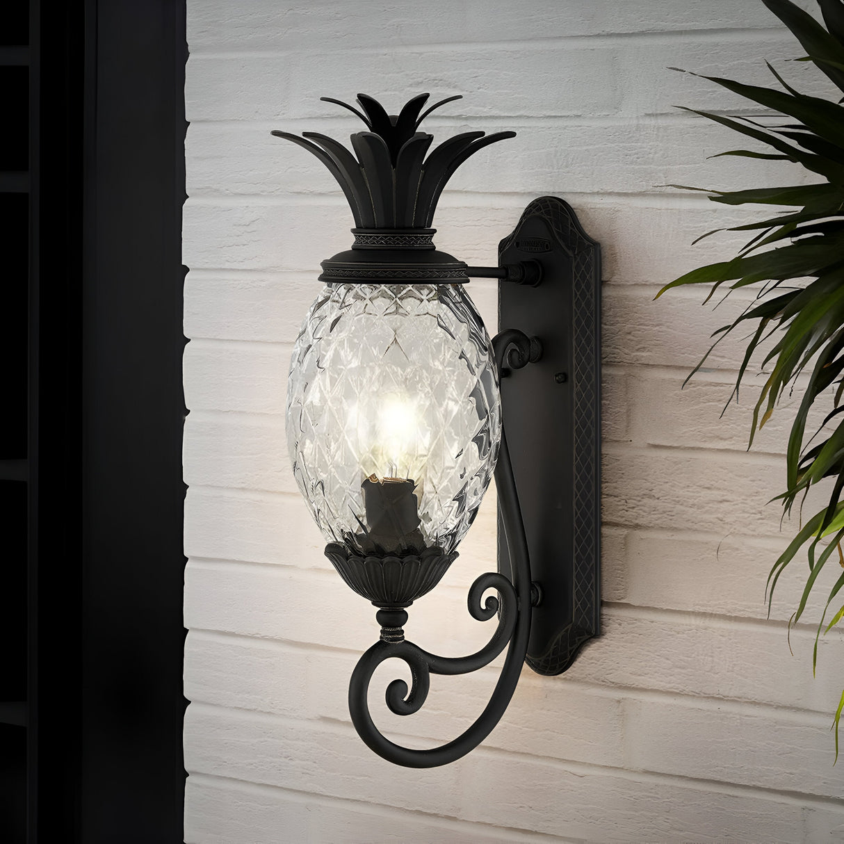 The Plantation Outdoor 1 Light Medium Wall Lantern in black, featuring a pineapple-shaped glass, radiates a warm glow against the textured white brick wall, providing an elegant touch to outdoor lighting. Lush green leaves are partially visible on the right, enhancing the inviting scene.