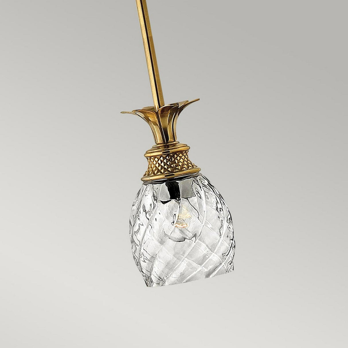 A close-up of the Plantation 1 Light Pendant in burnished brass, showcasing its elegant ceiling light fixture with a clear optic glass shade shaped like a pineapple. The background is a soft, pale gray.