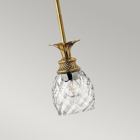 A close-up of the Plantation 1 Light Pendant in burnished brass, showcasing its elegant ceiling light fixture with a clear optic glass shade shaped like a pineapple. The background is a soft, pale gray.