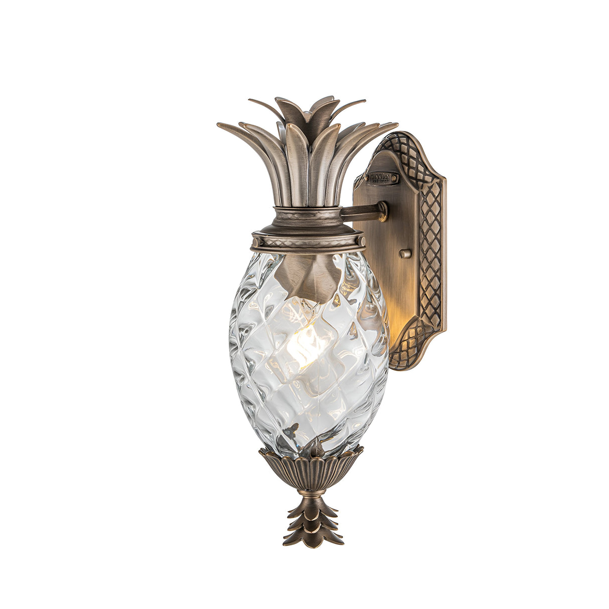 The Plantation Outdoor 1 Light Small Wall Lantern in burnished bronze is an elegant sconce designed in the shape of a pineapple, featuring a glass body. Its intricate metalwork adorned with a leaf motif complements the top and bottom, while light beautifully shimmers through the pineapple optic glass.