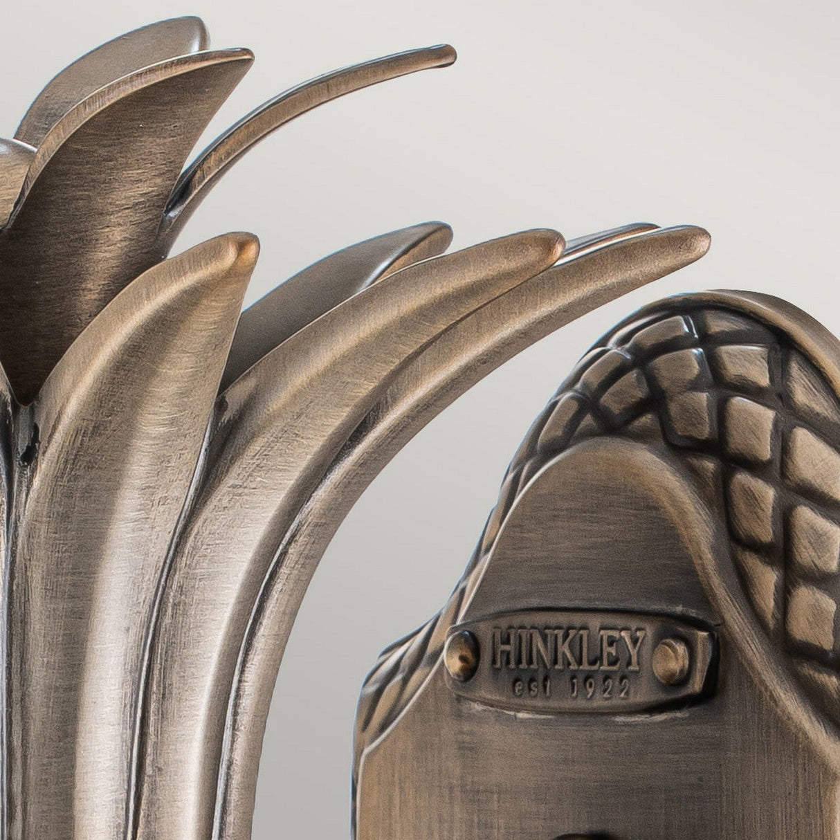 Close-up of the Plantation Outdoor 1 Light Small Wall Lantern in burnished bronze, showcasing abstract leaf-like designs on the left and a textured surface on the right. The word HINKLEY and est. 1922 are elegantly engraved, all set against a light gray background.