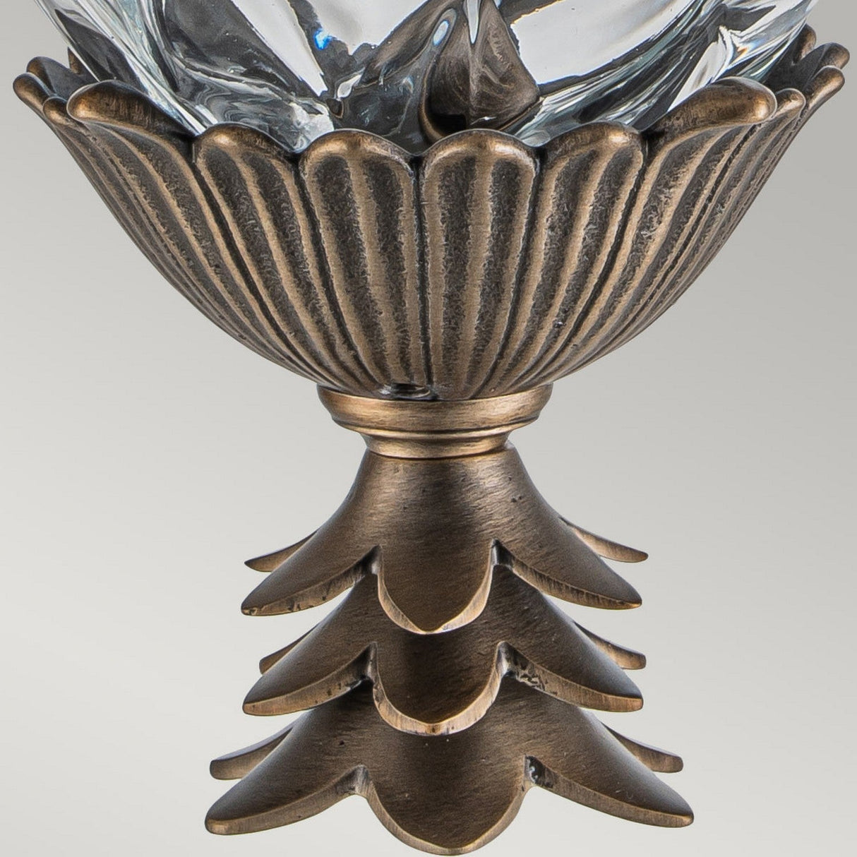 A decorative metallic sculpture designed as a wall lantern features a fluted, bowl-shaped top and a tiered, leaf-like base with a burnished bronze finish. The design is enhanced by an optic glass centerpiece resembling a pineapple, complete with faceted clear components, bringing elegance to any setting.