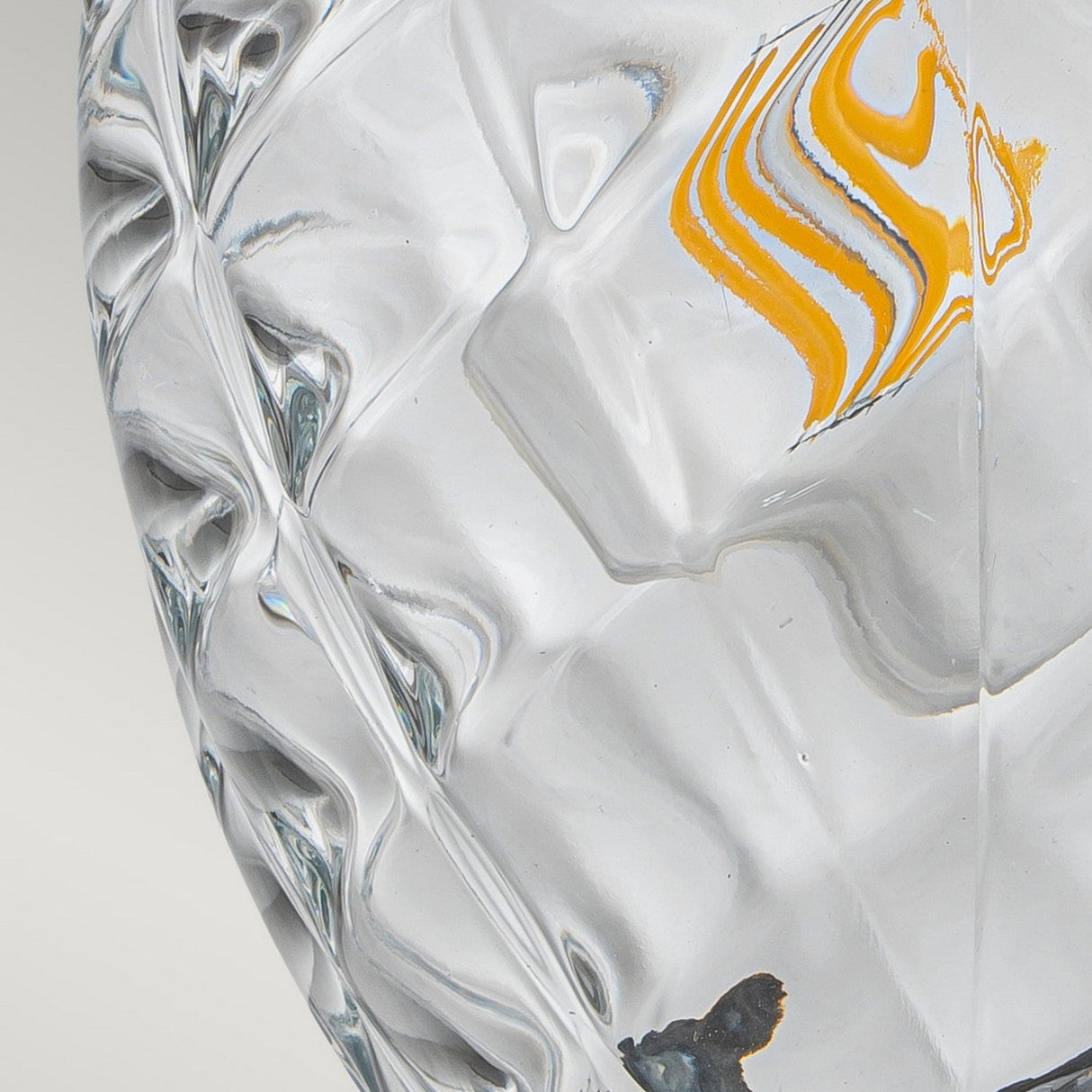 A close-up of the Plantation Outdoor 1 Light Small Wall Lantern in Burnished Bronze features intricate geometric patterns and swirls reminiscent of pineapple optic glass, with light reflections revealing subtle hints of orange and gray accents against its burnished bronze backdrop.