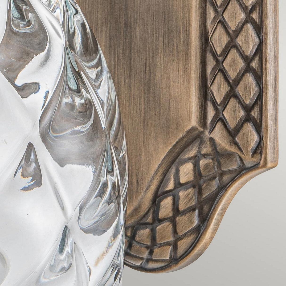 Close-up of the Plantation Outdoor 1 Light Small Wall Lantern, showcasing an intricate swirling pineapple optic glass design on the left and a textured, wood-like backplate with a lattice pattern on the right. The burnished bronze finish seamlessly combines modern and rustic elements.