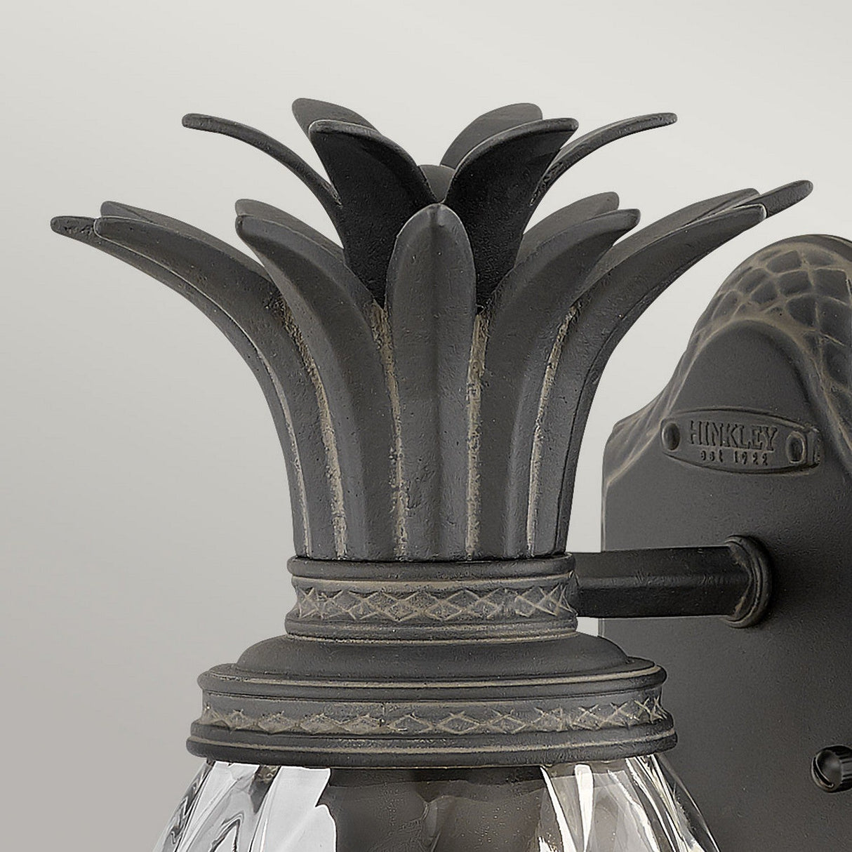Close-up of the Plantation Outdoor 1 Light Small Wall Lantern - Black. The design features a stylized black pineapple crown with intricate detailing and a sleek black finish, while the base showcases textured, pineapple-shaped glass.