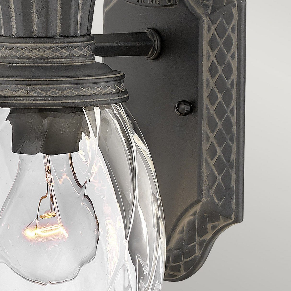 A close-up of the Plantation Outdoor 1 Light Small Wall Lantern - Black highlights its decorative backplate and pineapple-shaped glass globe surrounding a lit bulb. The fixture, featuring an elegant black finish, is mounted on a smooth, light gray wall.