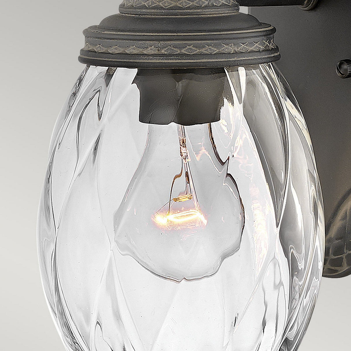 A detailed view of the Plantation Outdoor 1 Light Small Wall Lantern reveals its clear, textured, pineapple-shaped glass shade that showcases the glowing bulb inside, all supported by a sleek black metal base.