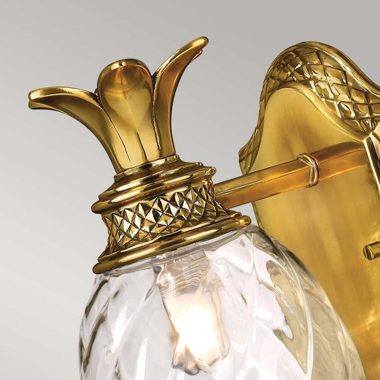 A close-up of the Plantation Outdoor 1 Light Wall Light – Burnished Brass reveals a fixture with a glass globe elegantly shaped like a pineapple. The gold-colored top and mount feature intricate detailing, enhancing the light's ornate appearance.