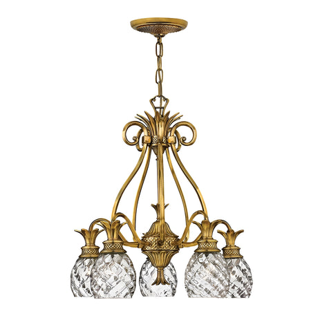 The Plantation 5 Light Chandelier - Burnished Brass is a vintage-style chandelier that showcases a burnished brass frame and five intricate crystal lampshades. Its design features scrolling metalwork adorned with decorative elements, including glass accents shaped like pineapples, and it is elegantly suspended by a chain.