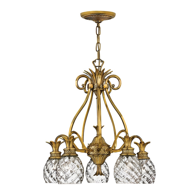 The Plantation 5 Light Chandelier - Burnished Brass is a vintage-style chandelier that showcases a burnished brass frame and five intricate crystal lampshades. Its design features scrolling metalwork adorned with decorative elements, including glass accents shaped like pineapples, and it is elegantly suspended by a chain.