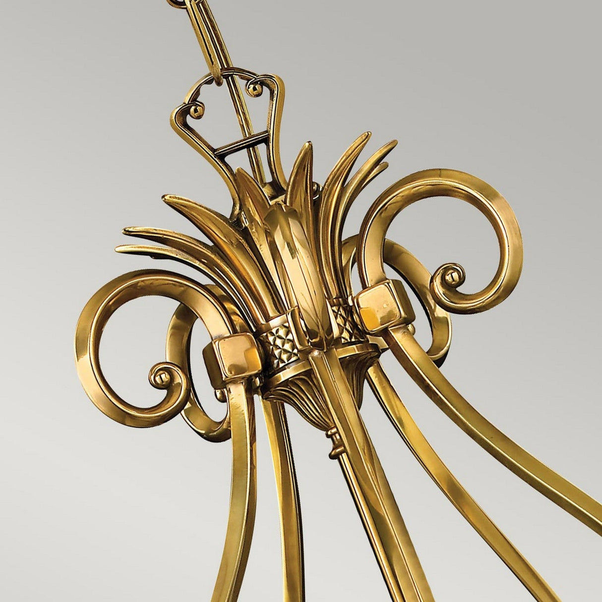 Close-up of the Plantation 5 Light Chandelier in burnished brass, showcasing its decorative gold-colored metal with intricate scrollwork and floral motifs. The curved elements of this elegant piece stand out beautifully against a light gray background, enhancing its ornate charm.