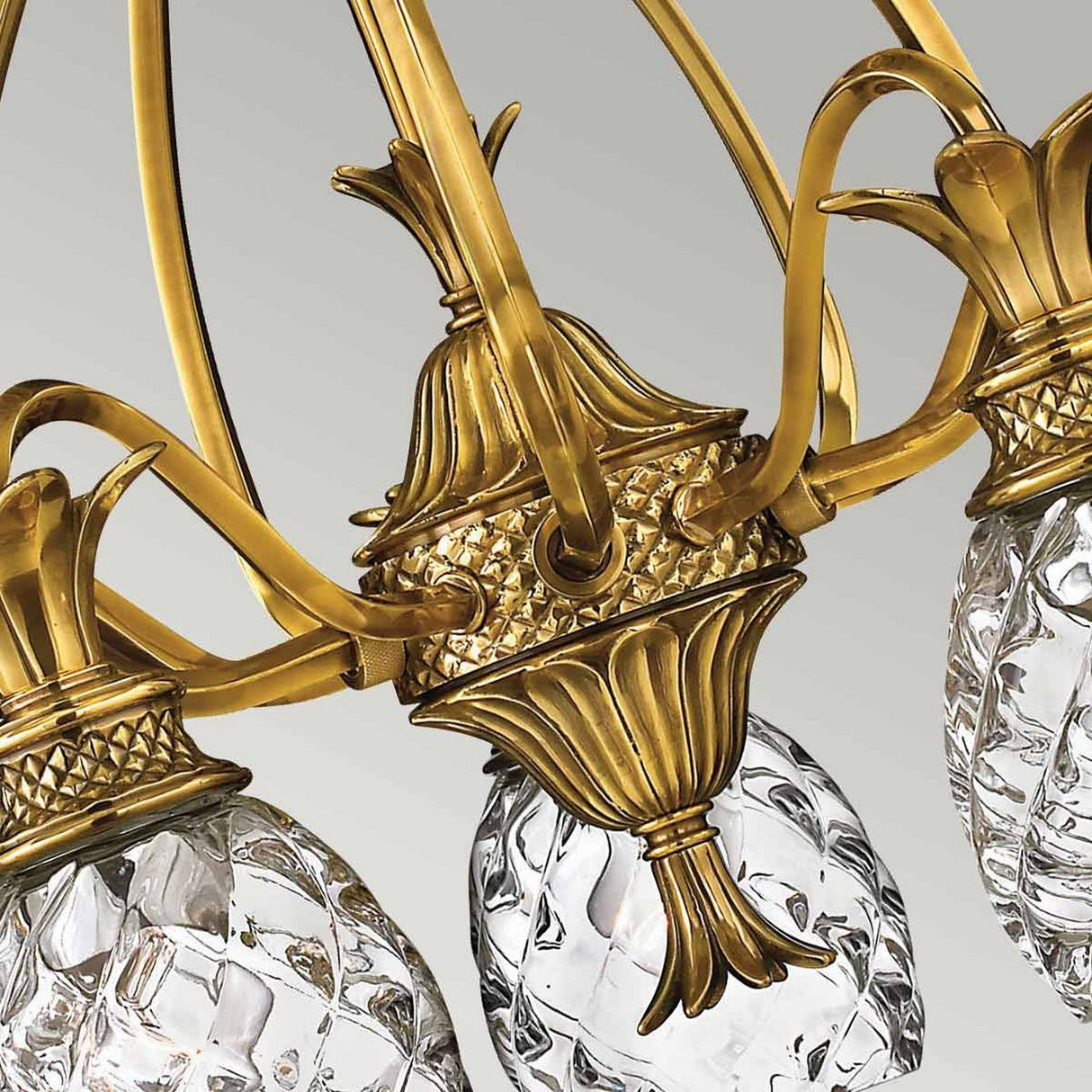 A close-up of the Plantation 5 Light Chandelier in burnished brass showcases its elaborate metalwork and clear crystal elements fashioned like pineapples. The design harmonizes intricate detailing with a luxurious, tropical style set against a neutral backdrop.