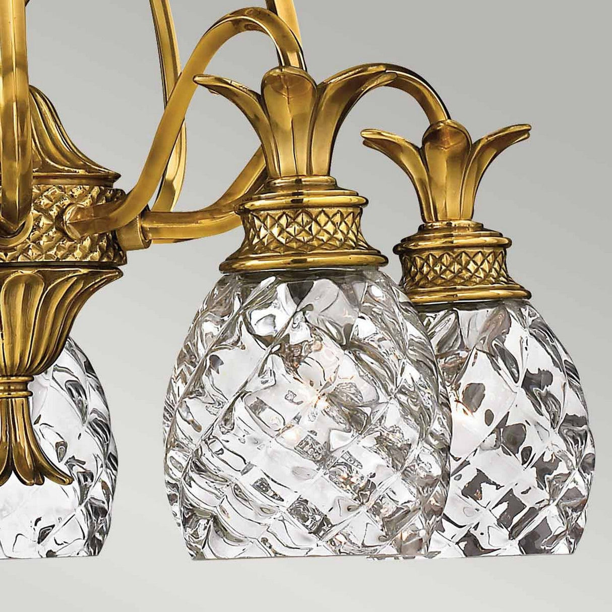 Close-up of the Plantation 5 Light Chandelier - Burnished Brass, featuring five pineapple-shaped glass pendants accented with burnished brass. The intricate glass design and metallic features lend it an elegant and tropical appearance.