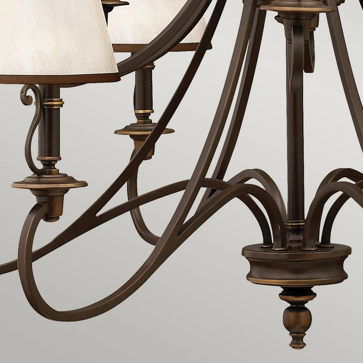 A close-up of the Plymouth 15 Light Chandelier in bronze, featuring ornate curving metalwork and vintage-inspired detailing with cream-colored lampshades, set against a neutral background to create an elegant ambiance.