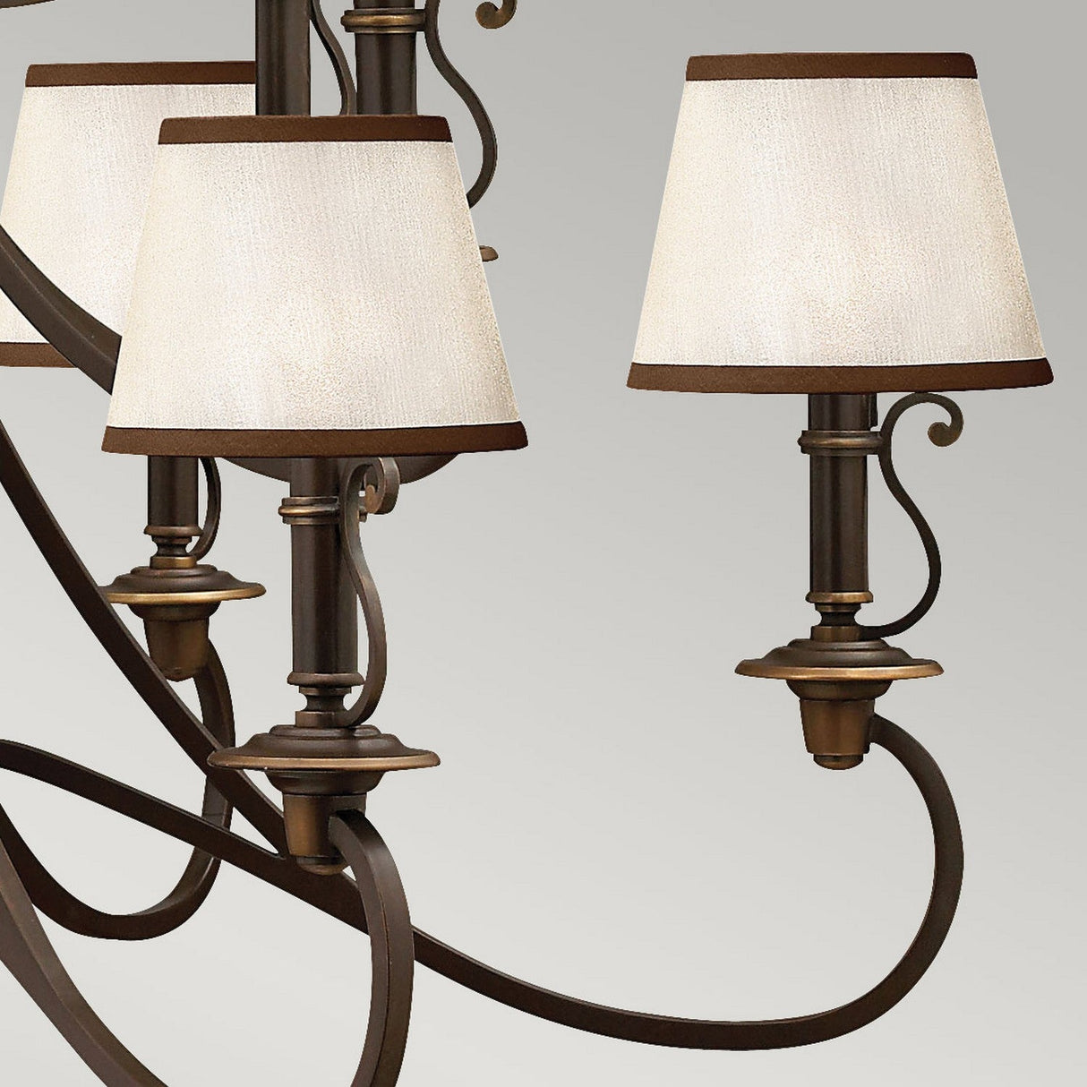 A close-up of the Plymouth 15 Light Chandelier - Bronze highlights its vintage-inspired elegance, featuring three slender arms adorned with beige fabric lampshades edged in dark trim. The design is defined by elegant curves and intricate scrollwork, standing out beautifully against a plain background.
