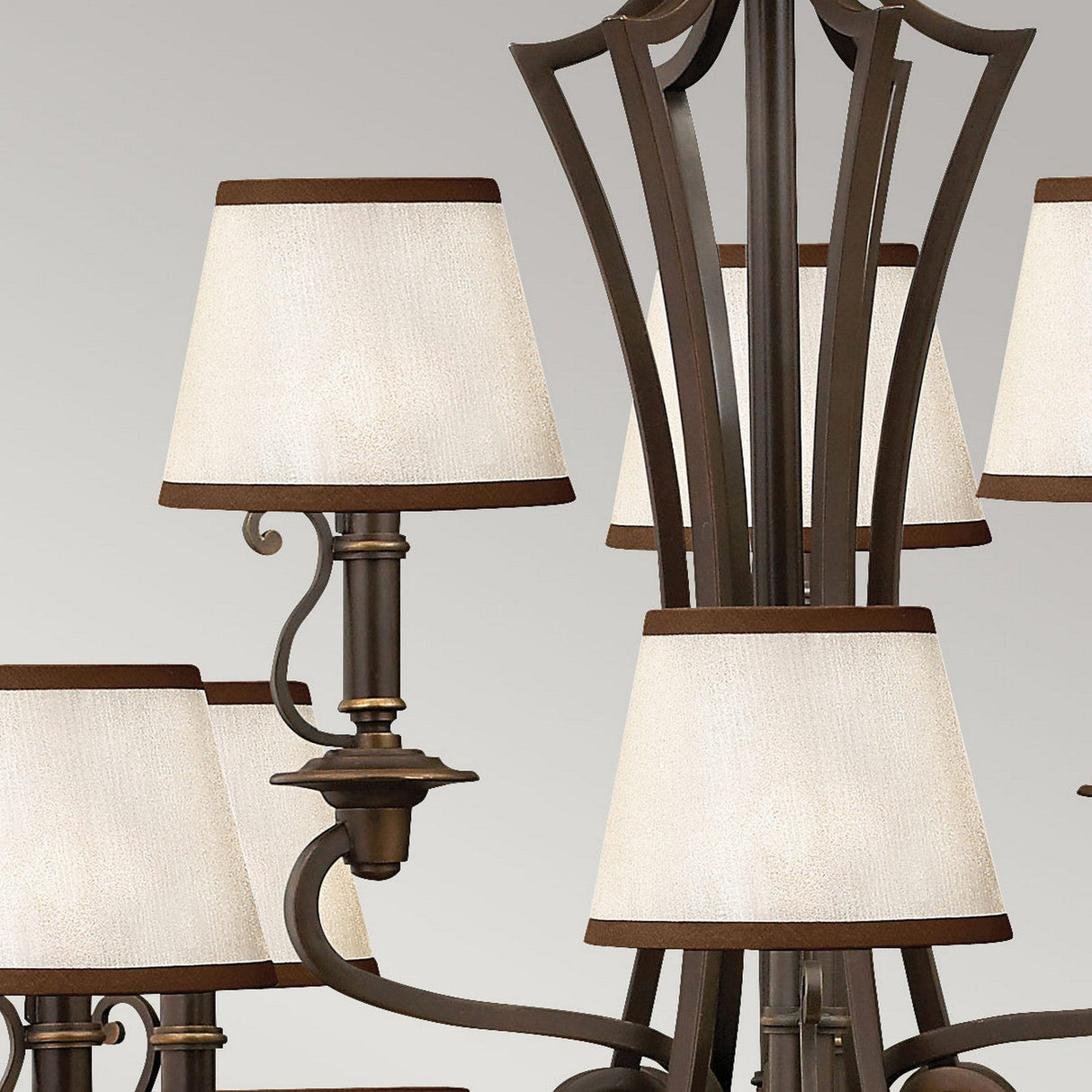 A close-up of the Plymouth 15 Light Chandelier in bronze, highlighting its vintage-inspired design with multiple cream-colored lampshades adorned with dark trim, supported by elegantly curved arms against a light gray background.
