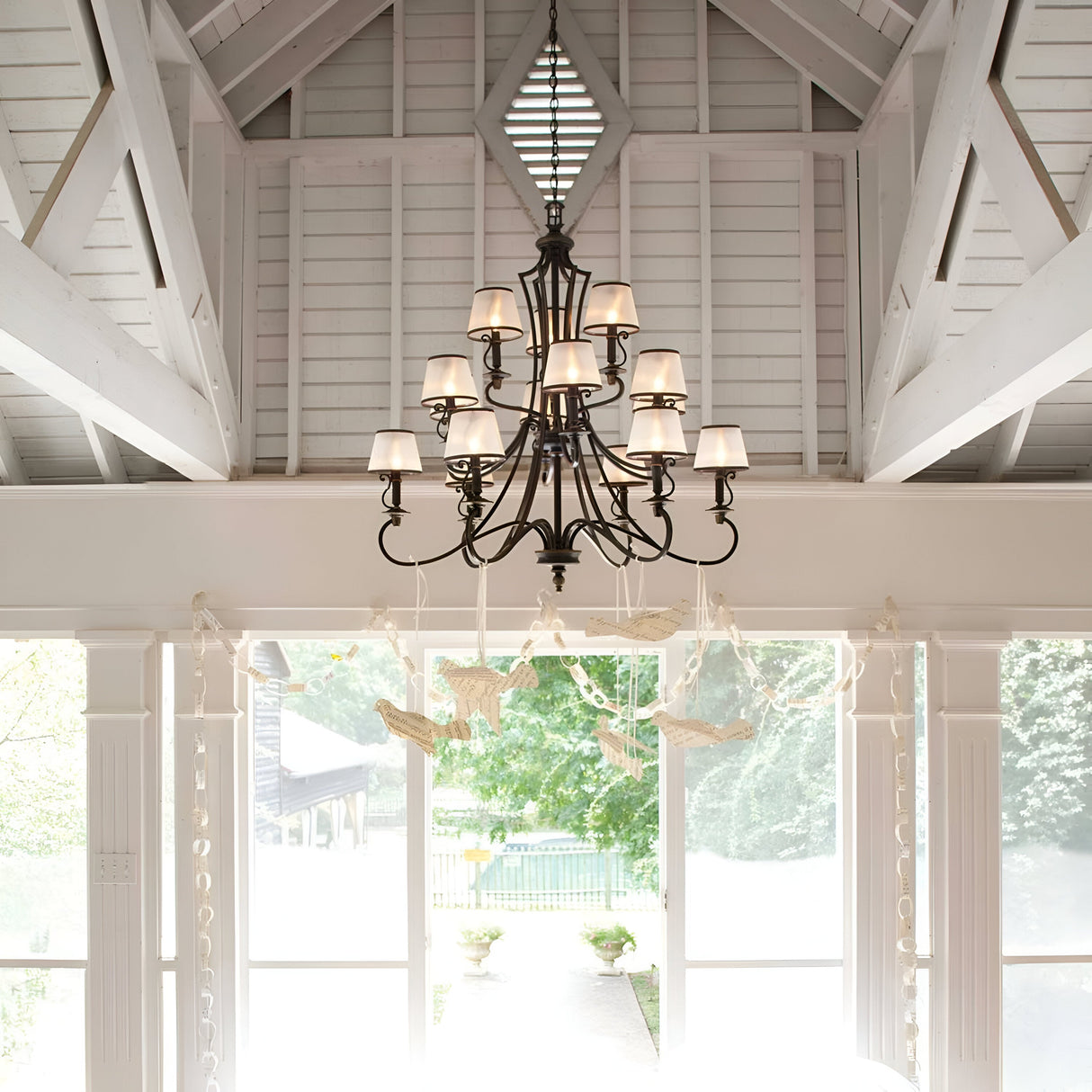 A vintage-inspired Plymouth 15 Light Chandelier - Bronze with white lampshades hangs from a high, vaulted wooden ceiling. Below, large windows offer a view of a sunlit garden path lined with greenery, creating an elegant and airy interior space.