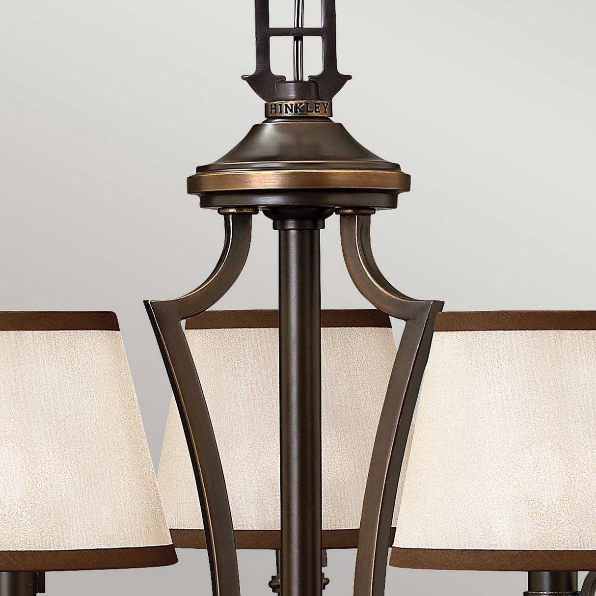 Close-up of the Plymouth 3 Light Chandelier - Bronze with a detailed central design. This elegant lighting piece features three beige fabric lampshades, offering a classic and sophisticated ambiance. The chandelier is accented by a dark, antique finish for a vintage-inspired look.