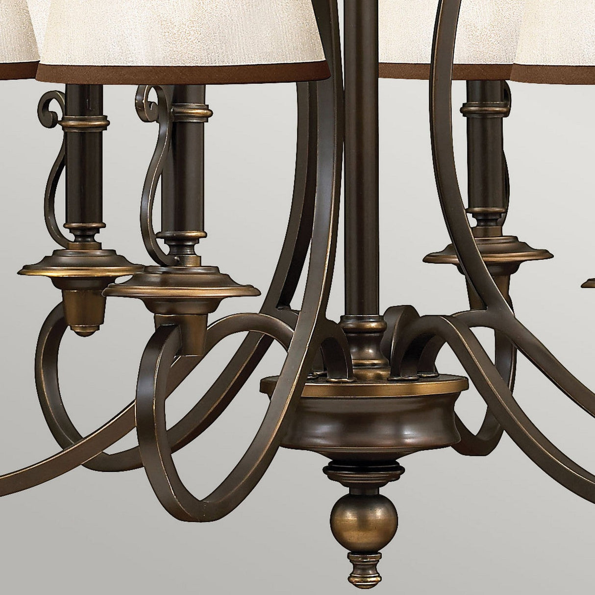 A close-up of the Plymouth 6 Light Chandelier - Bronze reveals its ornate curved arms and cream fabric shades, showcasing intricate scrolling metalwork that exudes vintage charm against a light gray background.