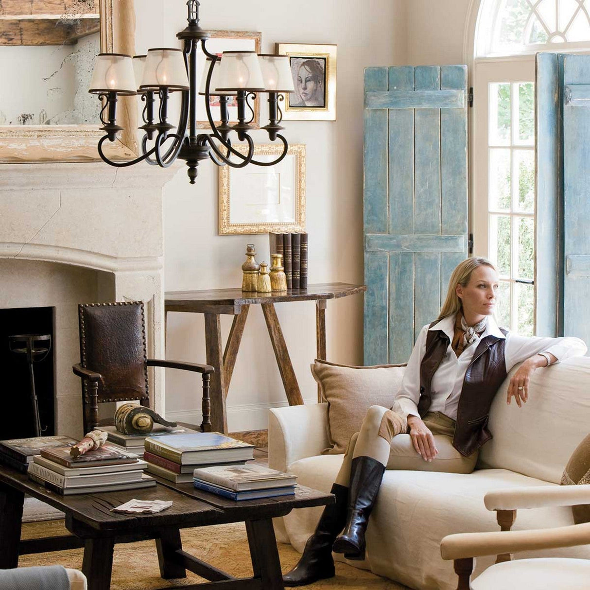 A person relaxes on a white sofa in a cozy living room filled with vintage charm. The rustic decor features a wooden table, books, and a fireplace. A Plymouth 6 Light Chandelier in bronze casts light over framed pictures and turquoise shutters by the bright window.
