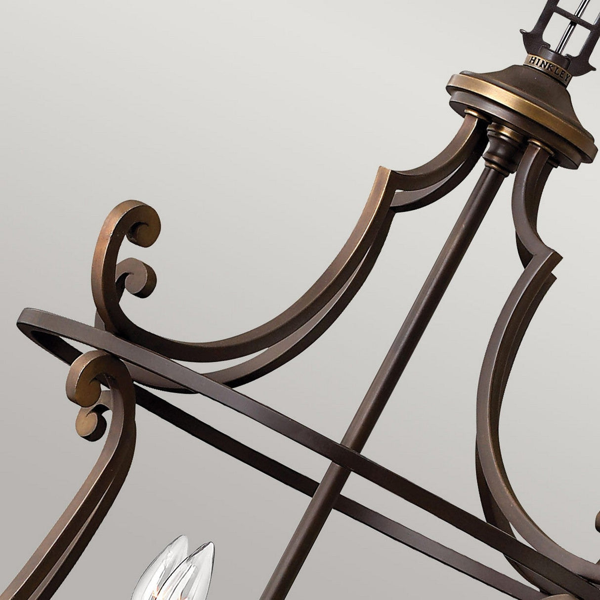 Close-up of the Plymouth 8 Light Chandelier - Bronze, showcasing its ornate metal frame with a bronze finish. The chandelier features elegant, curved designs and decorative scrollwork reminiscent of vintage candle sleeves. Its structure includes intersecting bars and highlights a visible light fixture at the top against a plain background.