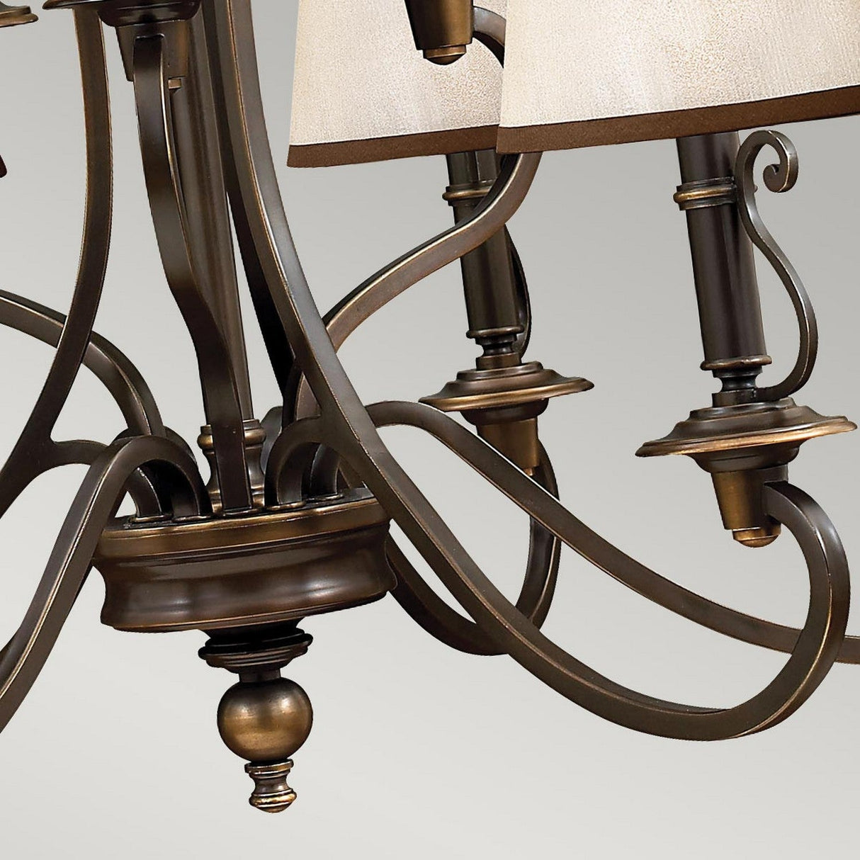 Captured in a close-up image is the sophisticated Plymouth 9 Light Chandelier - Bronze. The photograph emphasizes its gracefully curved arms decorated with vintage-inspired candle sleeves and intricate metalwork, all supporting beige lamp shades. The chandelier's design offers a classic and sophisticated flair against a neutral backdrop.