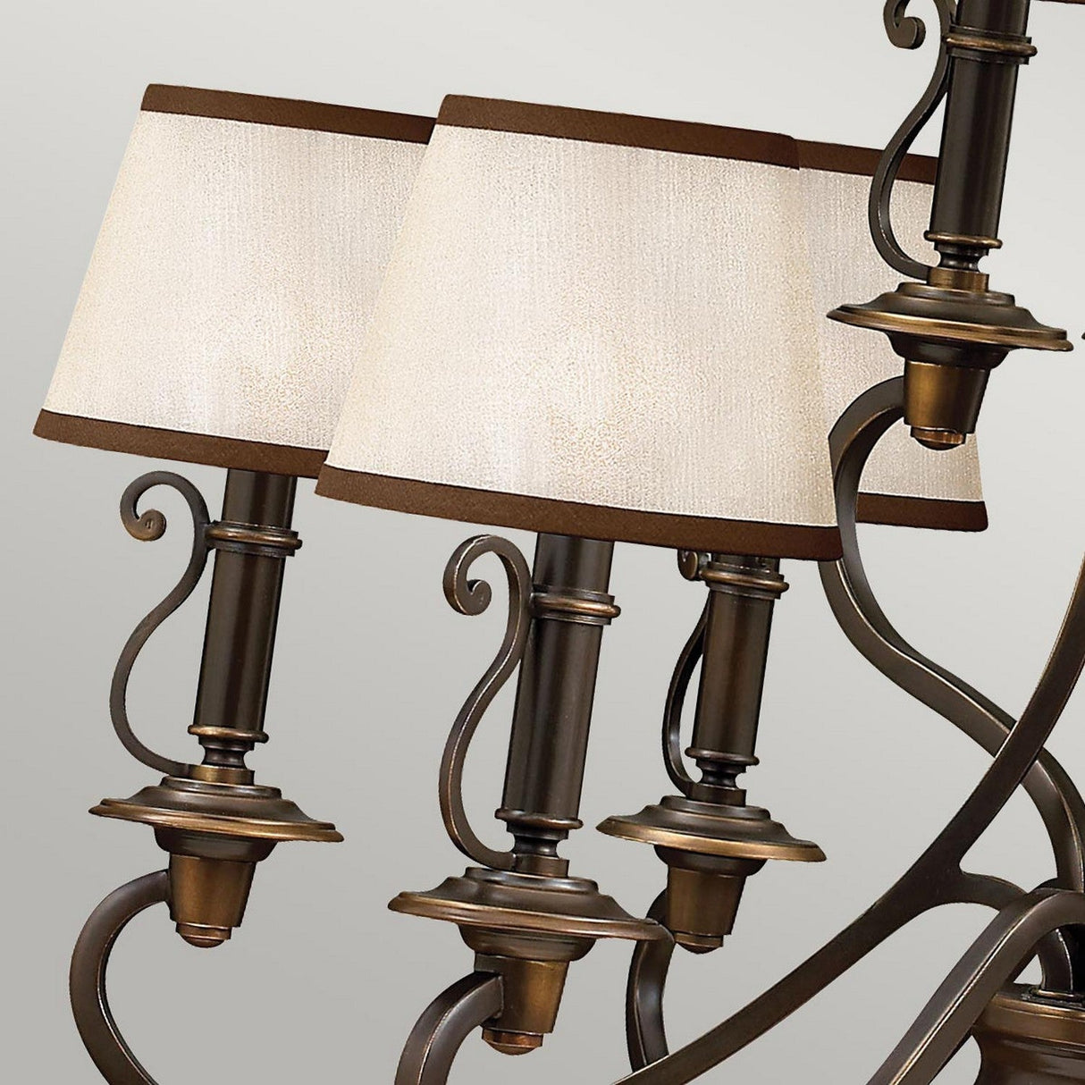 Close-up of the Plymouth 9 Light Chandelier - Bronze, showcasing three ornate bronze arms and cream-colored fabric lampshades. This chandelier, finished in Old Bronze, includes intricate scrollwork and vintage-inspired candle sleeves, adding an elegant touch against a neutral background.